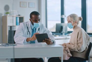 African American Doctor in Protective Mask is Reading Medical History of Senior Female Patient During Consultation in a Health Clinic. Physician Using Tablet Computer in Hospital Office.